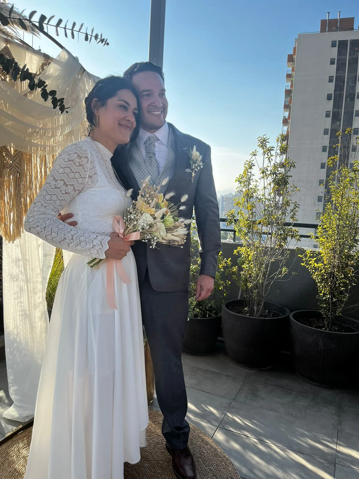 Los novios sonriendo en una terraza con luz de tarde, junto a un arco boho
